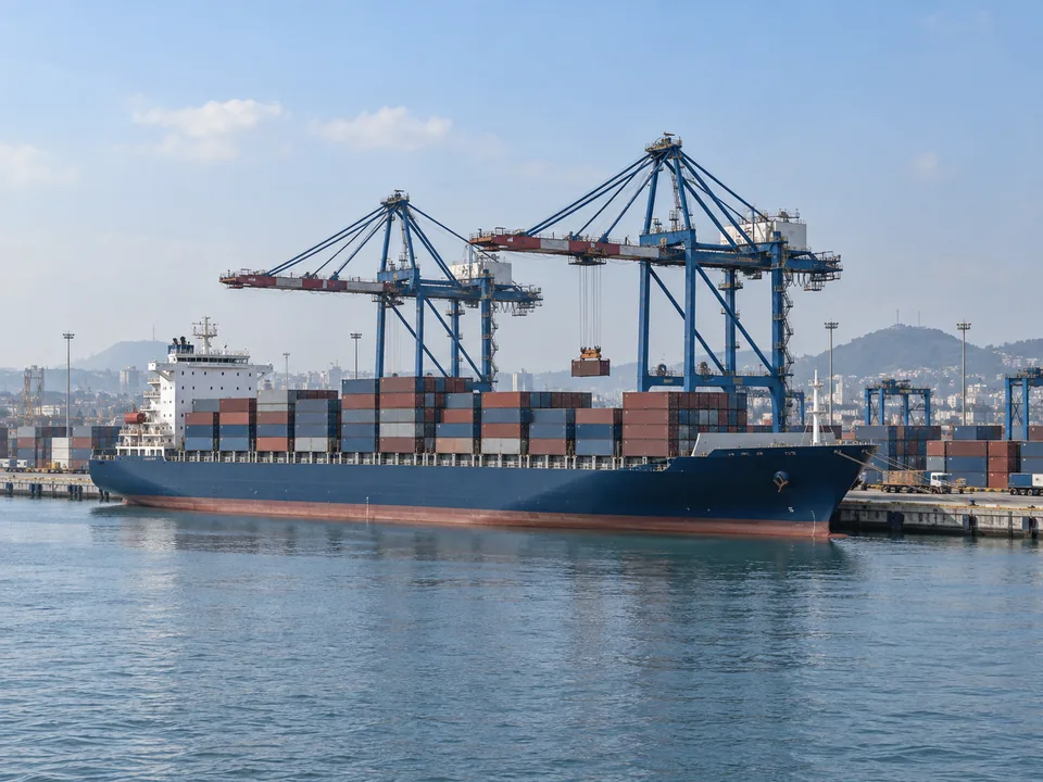 Container vessel moored beside quay cranes with harbour water in the foreground.